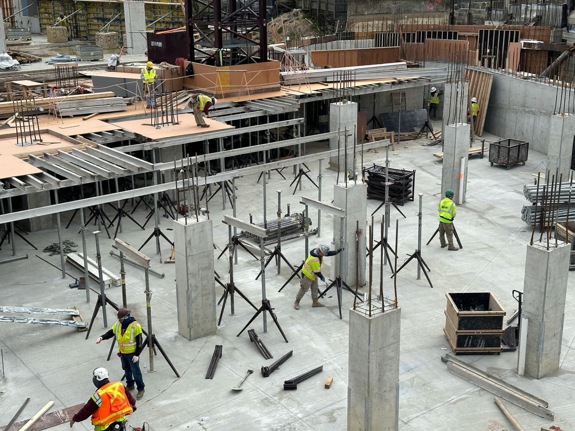 Construction workers in high-visibility vests and hard hats working on a concrete foundation slab with rebar and temporary supports.