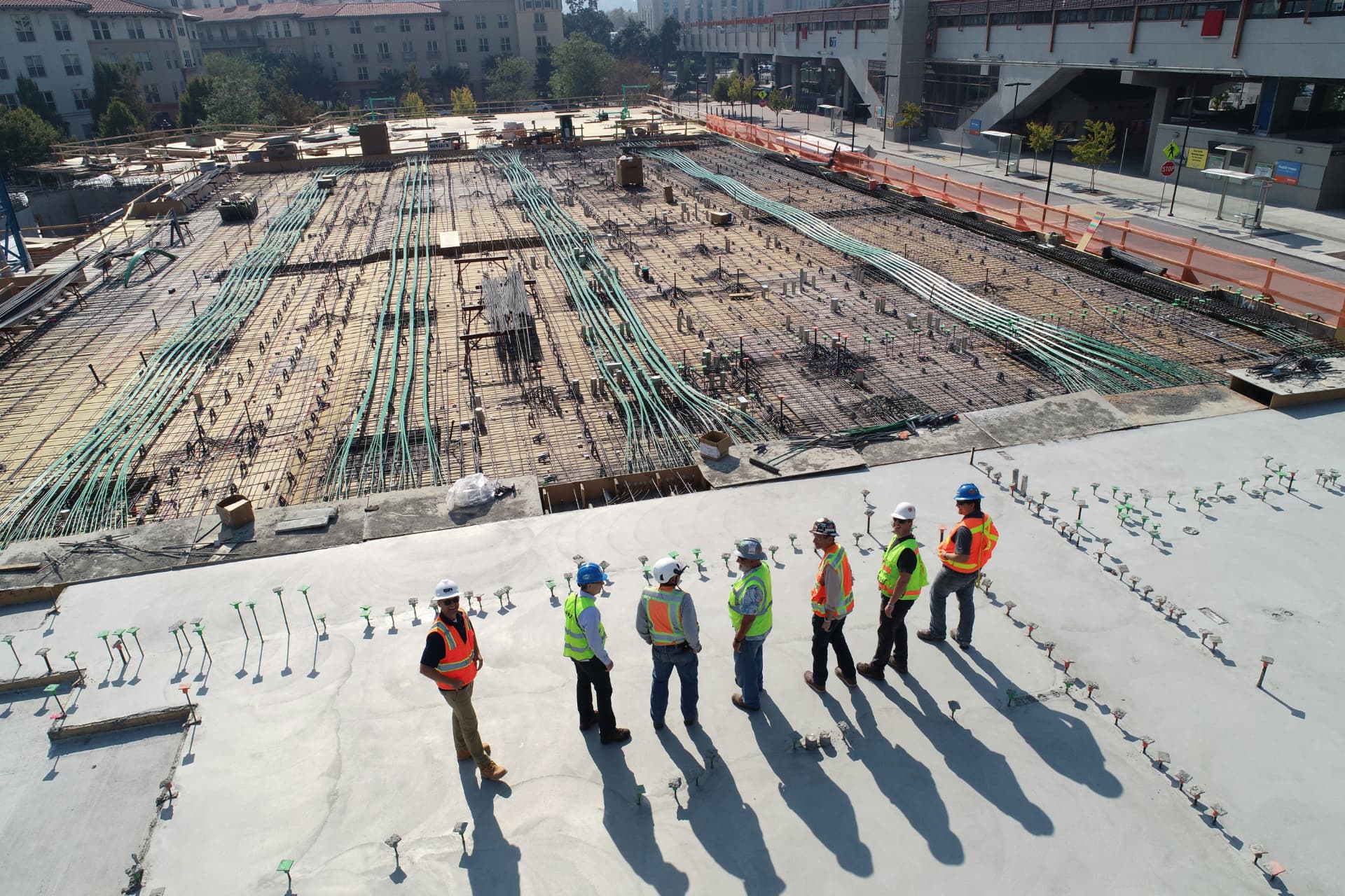 A team of concrete contractors standing on a freshly poured slab, reviewing a large rebar and post-tensioning layout at a commercial construction site.